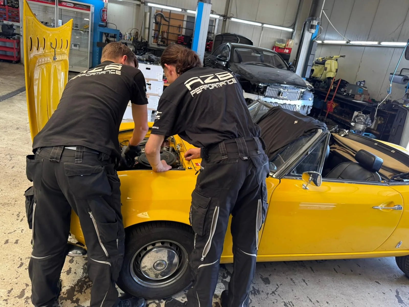 Zwei Mechaniker in AZB-Performance-Shirts arbeiten gemeinsam am offenen Motorraum eines gelben Fiat Spider in der Werkstatthalle